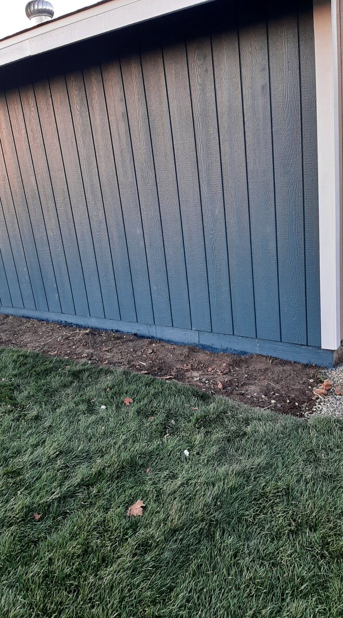 Blue-gray wooden shed wall beside a grassy lawn with a narrow strip of dirt at the base