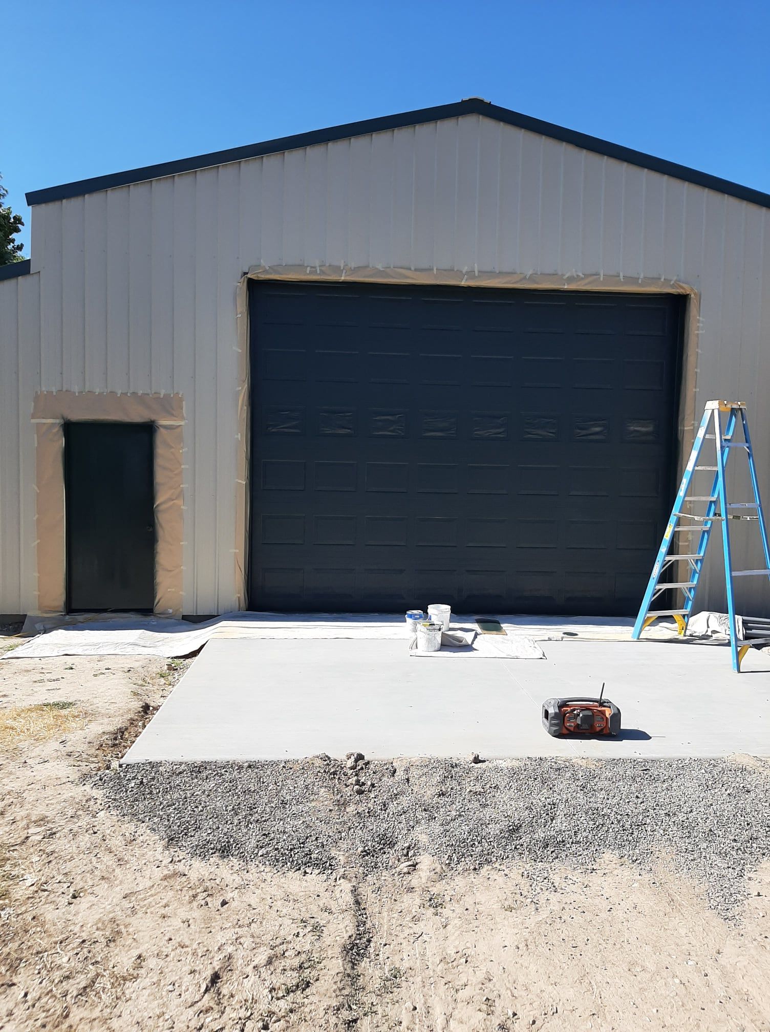 Small metal building with a large black garage door, open side door, and a ladder on a concrete driveway.