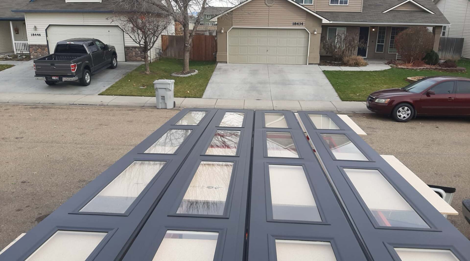 Gray folding doors laid across a driveway, with suburban houses and parked cars in the background.