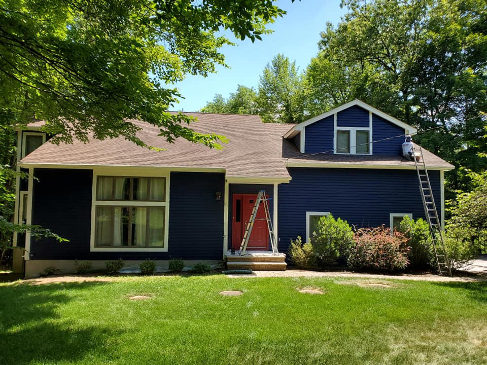 Blue house with red front door, white-trimmed windows, and a green lawn under trees.