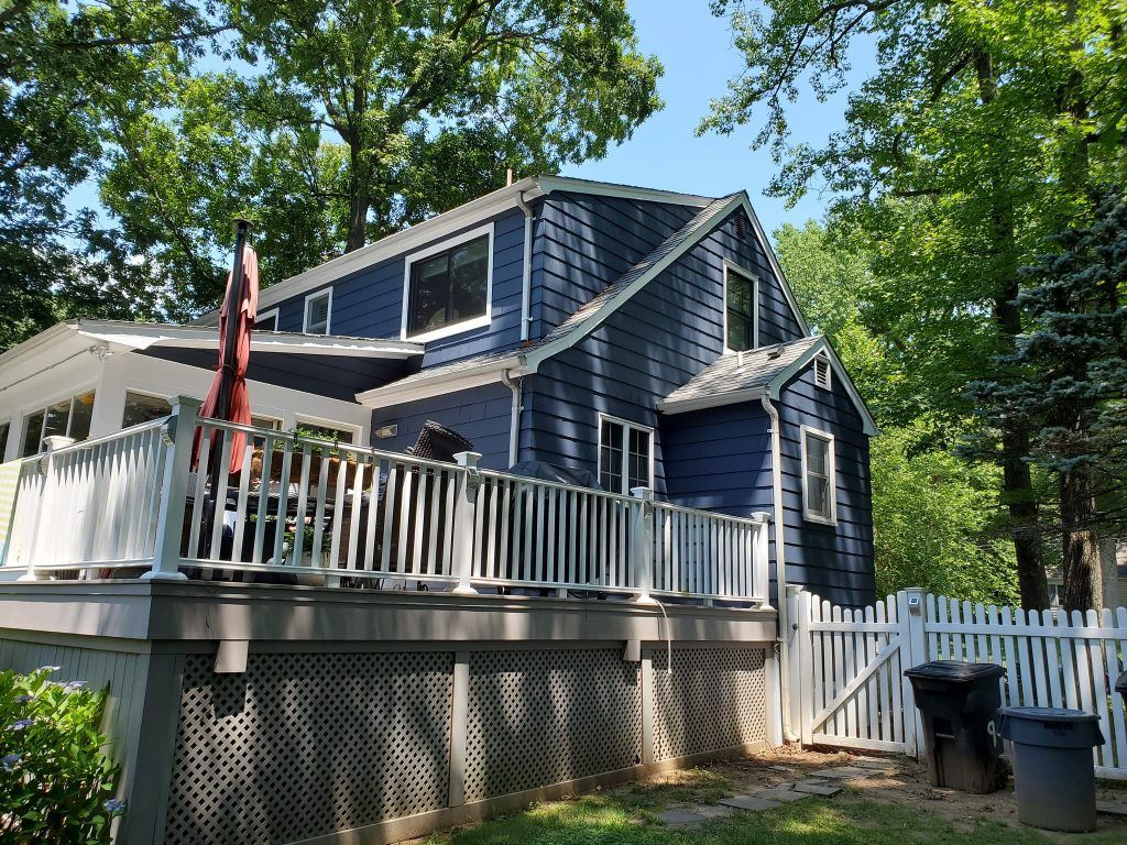 Blue house with white porch and deck, surrounded by trees and a white picket fence.