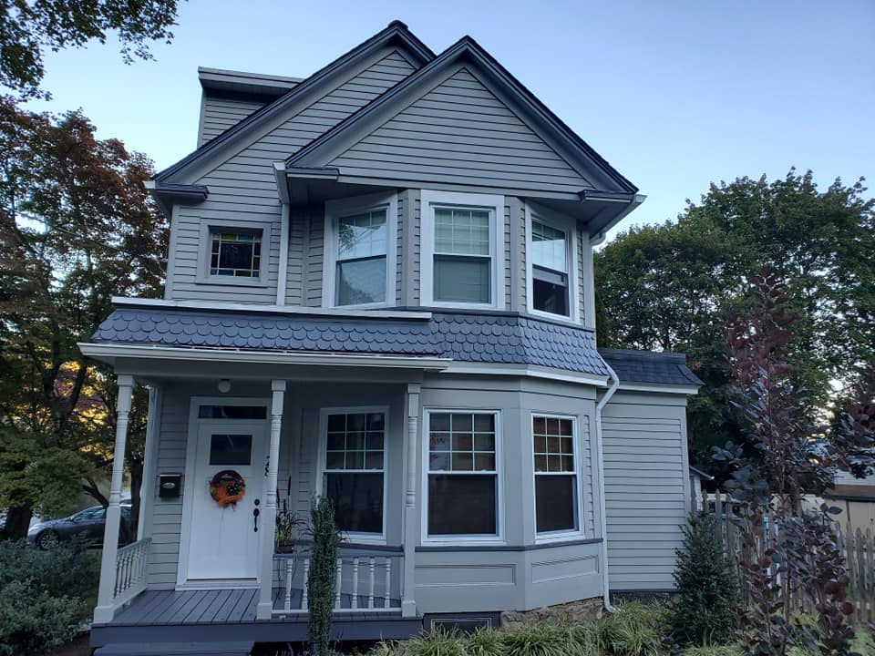 Gray two-story Victorian house with white trim and a small front porch, framed by trees at dusk