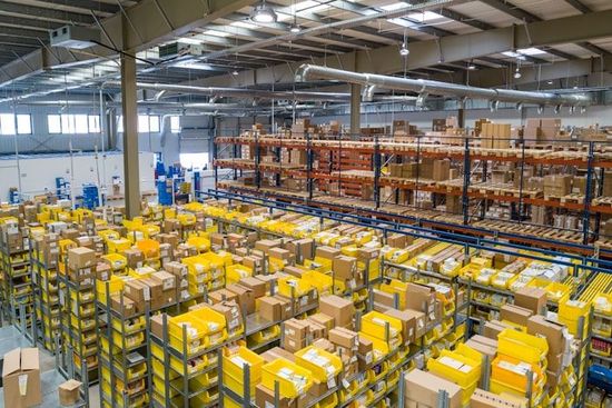 Groot magazijn vol gele dozen en stellingen onder fel industrieel licht.