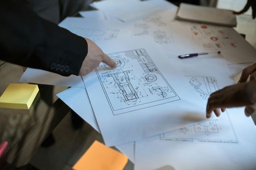 Handen wijzen naar architectuurtekeningen die over een tafel verspreid liggen, met plakbriefjes en een pen.