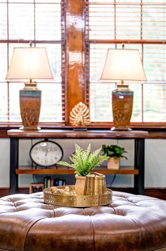 A living room with a ottoman , lamps , a clock and a plant on it.