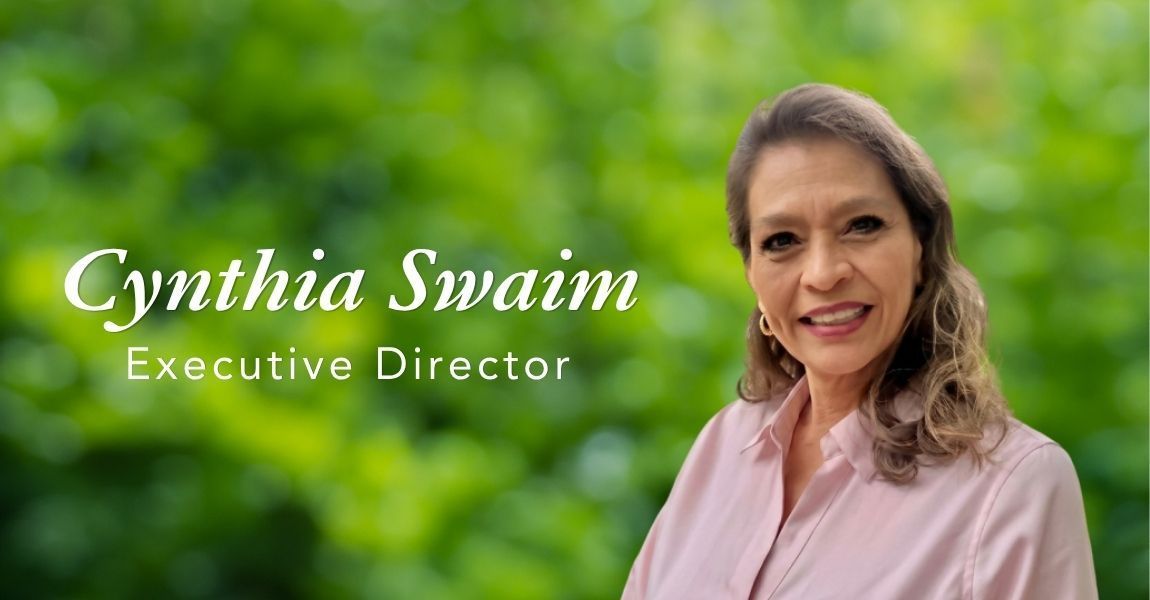 Cynthia Swain, Executive Director, smiling in a pink blouse against a green blurred background.
