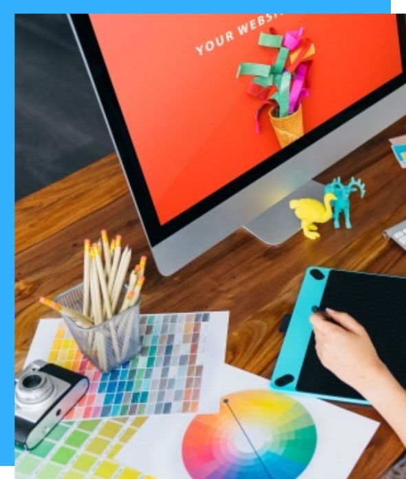 A desktop setup with a computer displaying website mockups, art supplies, and a graphic designer using a tablet.