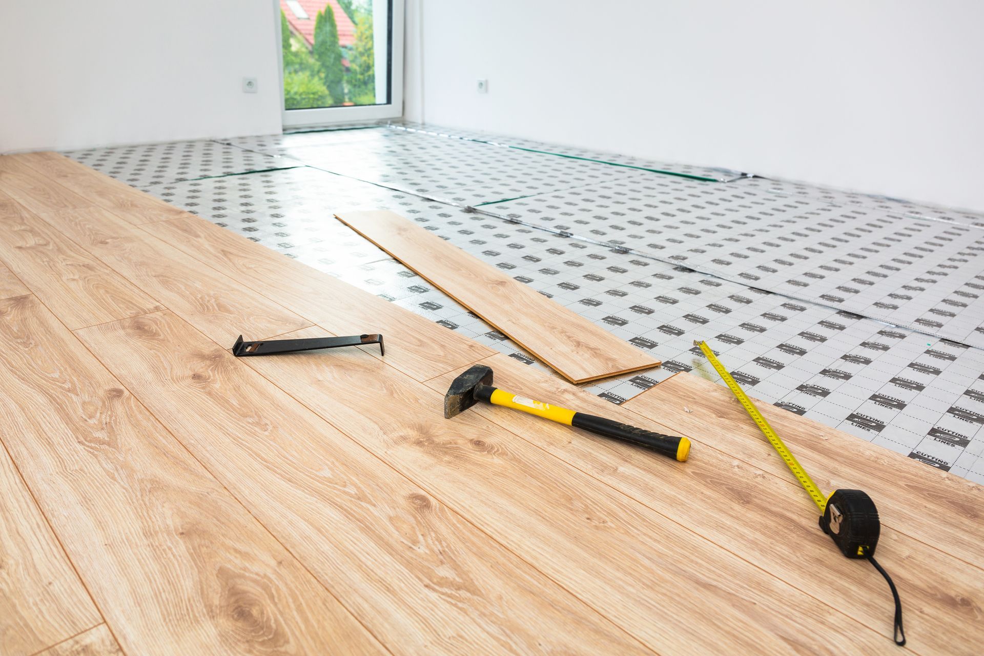 Installing laminate flooring in a room. Tools (hammer, tape measure) and planks are visible on a padded underlayment.