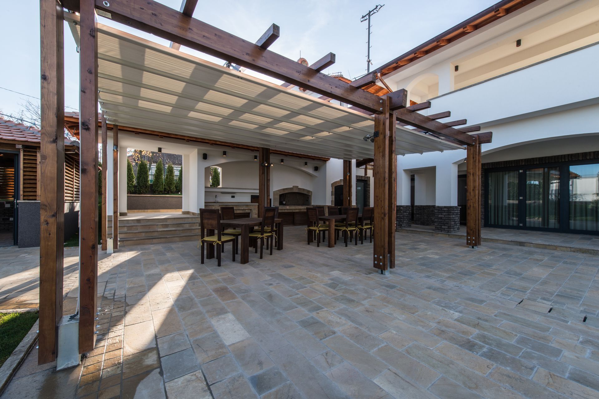 Covered outdoor patio with stone flooring, wooden pergola, and dining tables beside a modern house Covered outdoor patio with stone flooring, wooden pergola, and dining tables beside a modern house