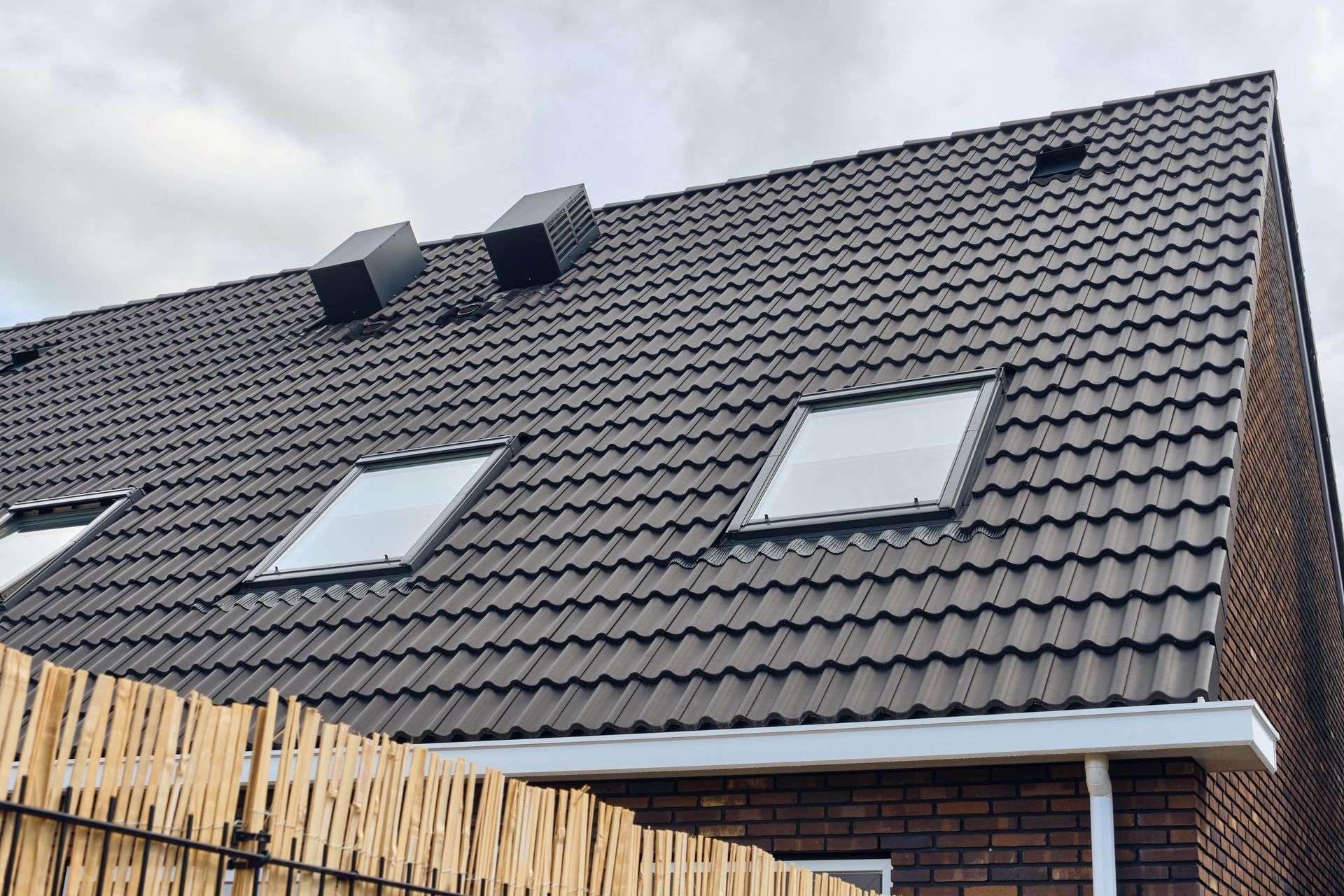 Dark gray tiled house roof with two skylights and two roof vents above a brick wall and fence Dark gray tiled house roof with two skylights and two roof vents above a brick wall and fence