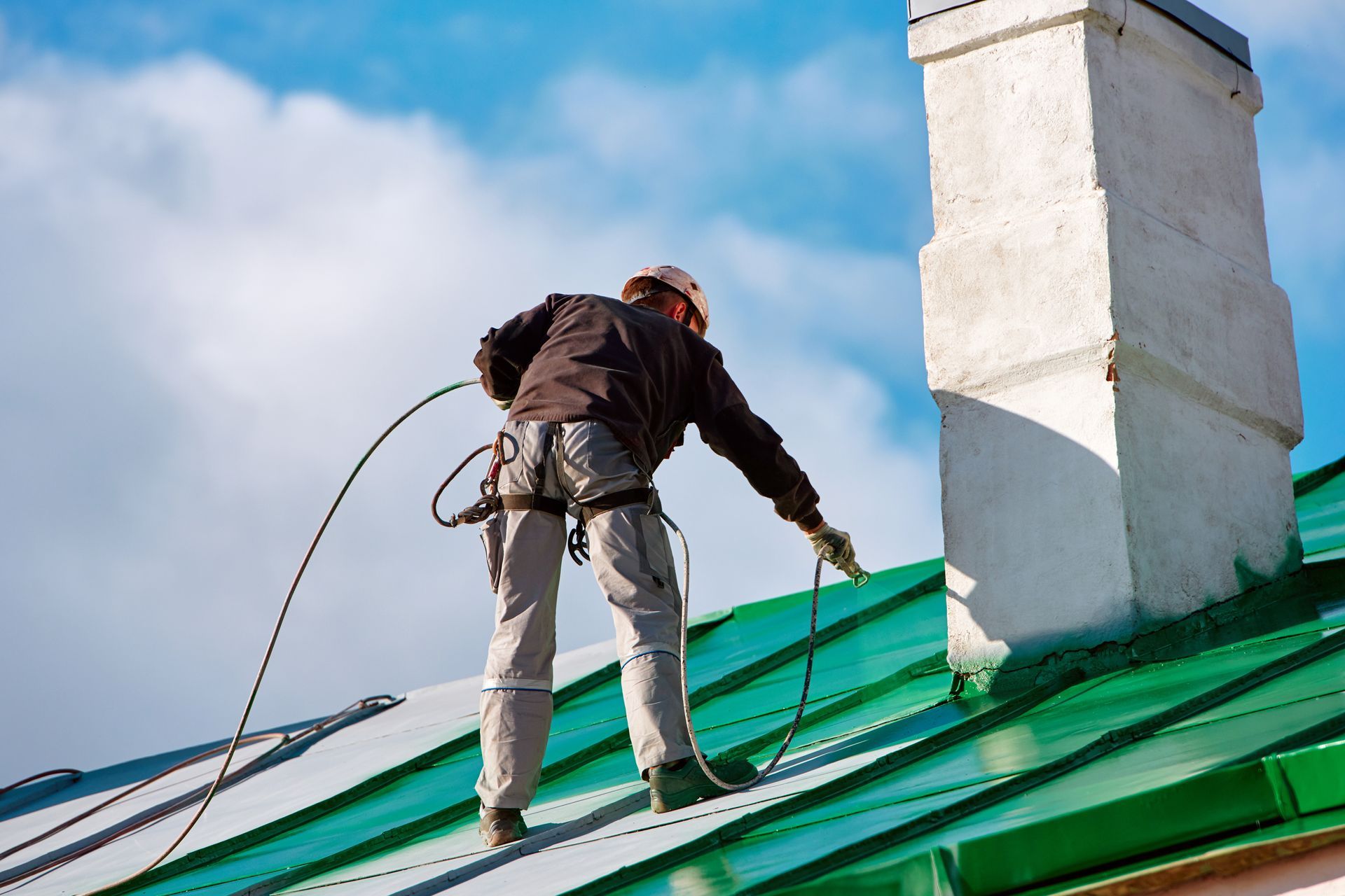 Worker on a green metal roof using a harness near a white chimney under a blue sky Worker on a green metal roof using a harness near a white chimney under a blue sky