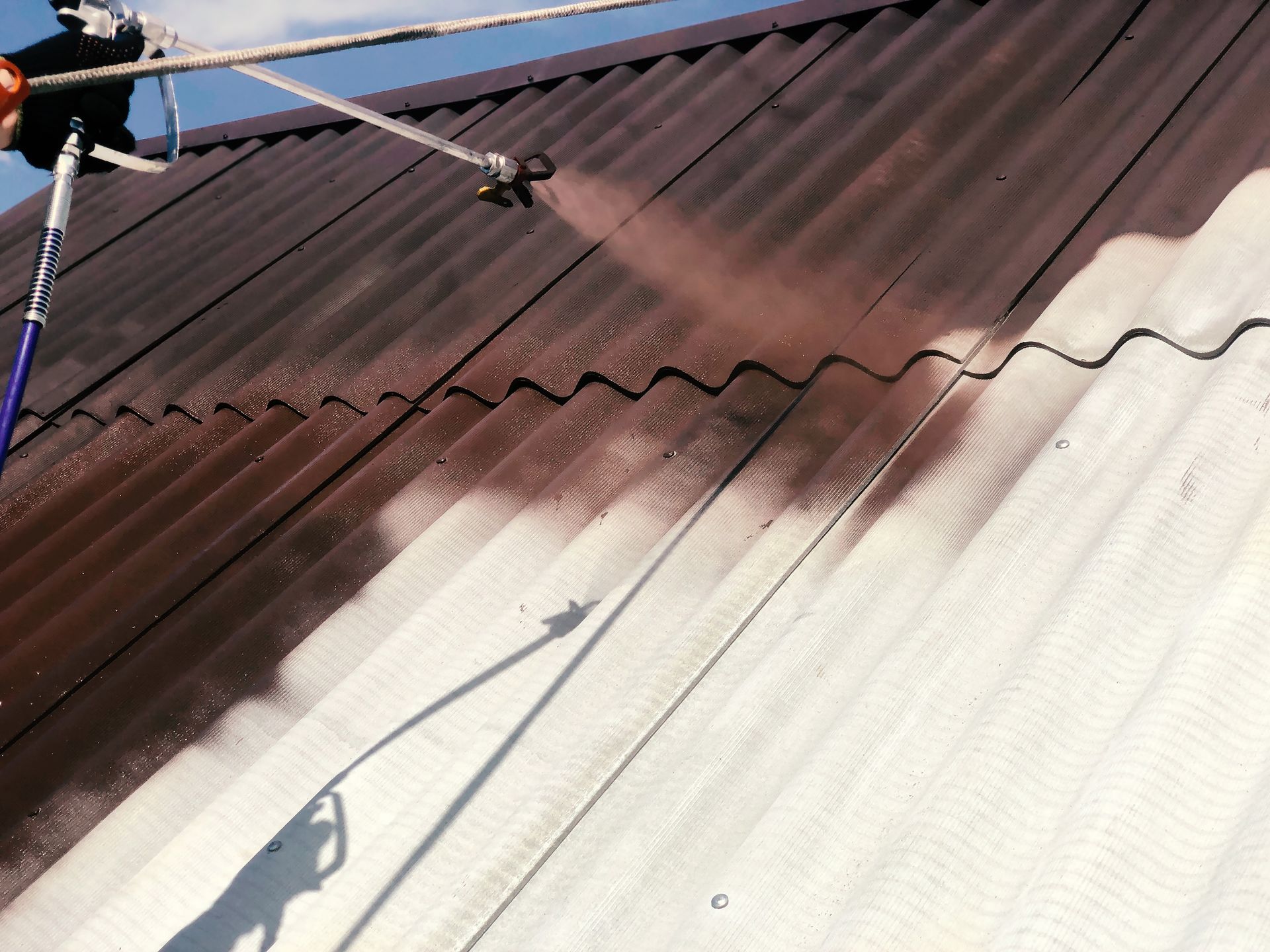 Worker pressure-washing a corrugated metal roof, half cleaned from dark brown to white.