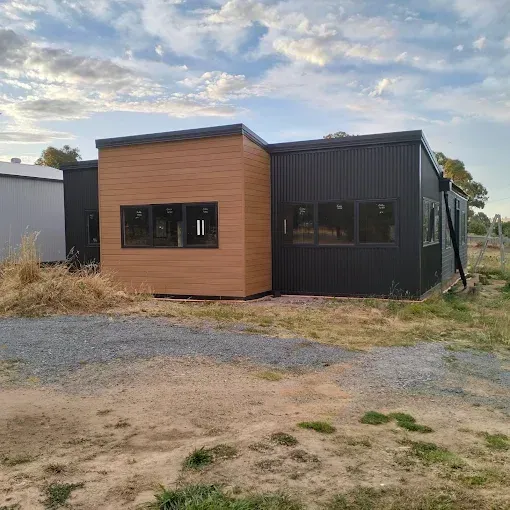 Small modern modular building with wood and black siding, sitting on a gravel lot under a cloudy sky Small modern modular building with wood and black siding, sitting on a gravel lot under a cloudy sky