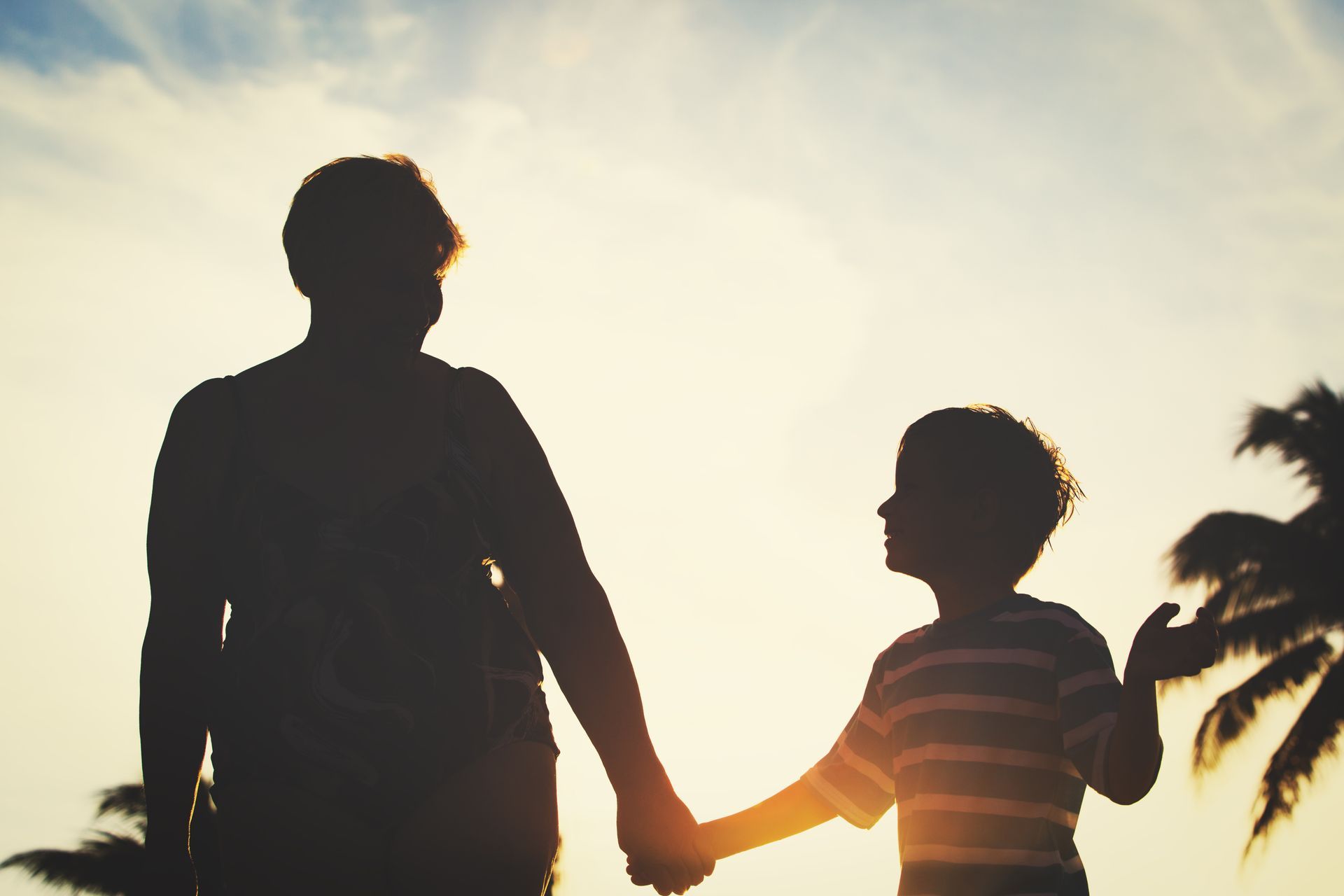 Silhouette of a person holding a child's hand. They walk toward the bright sun. Palm trees in the background.