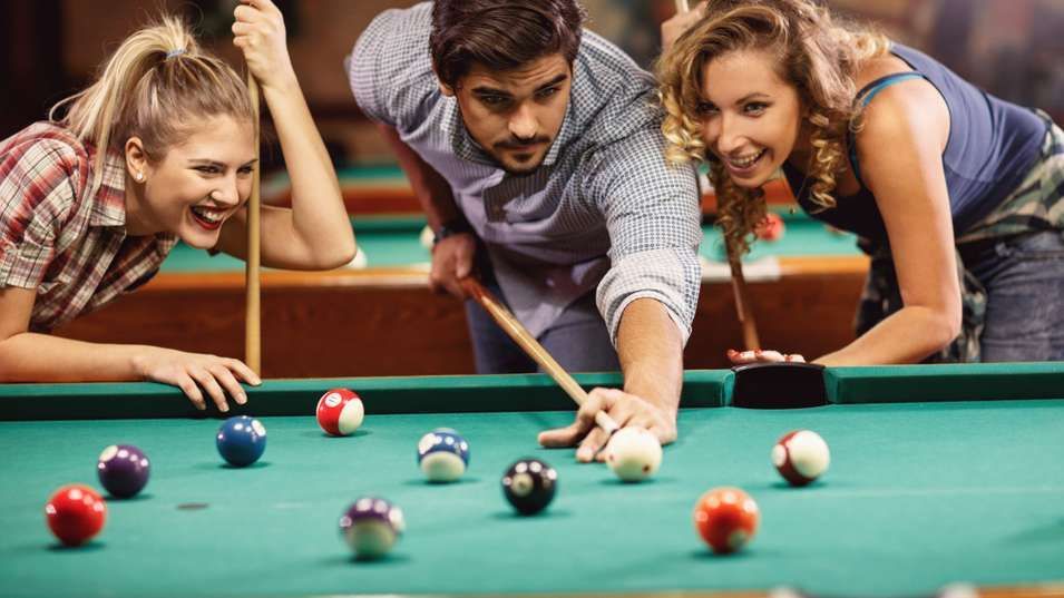 Three friends playing pool, smiling. Man lining up a shot; two women watching. Green table.