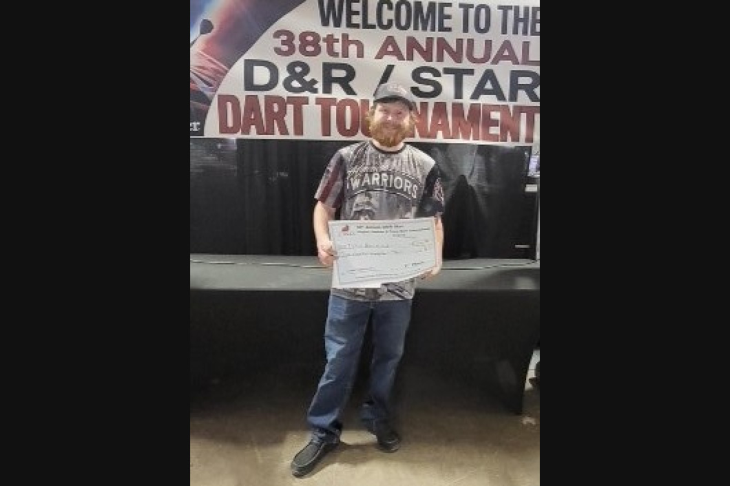 Person holding certificate, standing in front of a banner for a dart tournament.