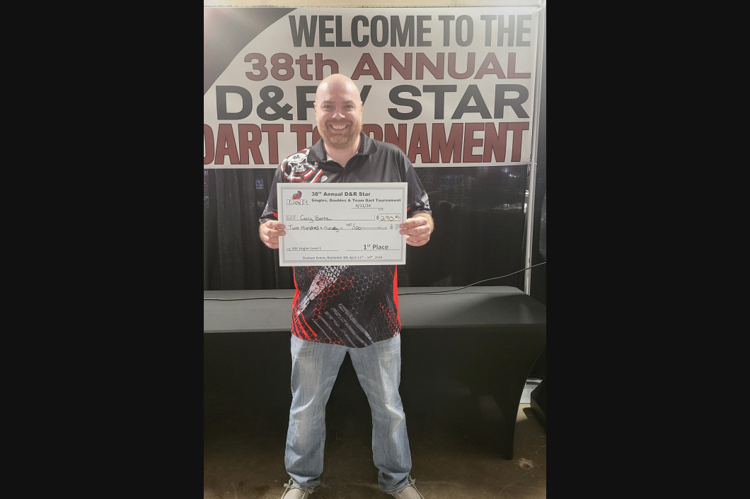 Man holding a check at a dart tournament. He's smiling, standing in front of a banner that reads 