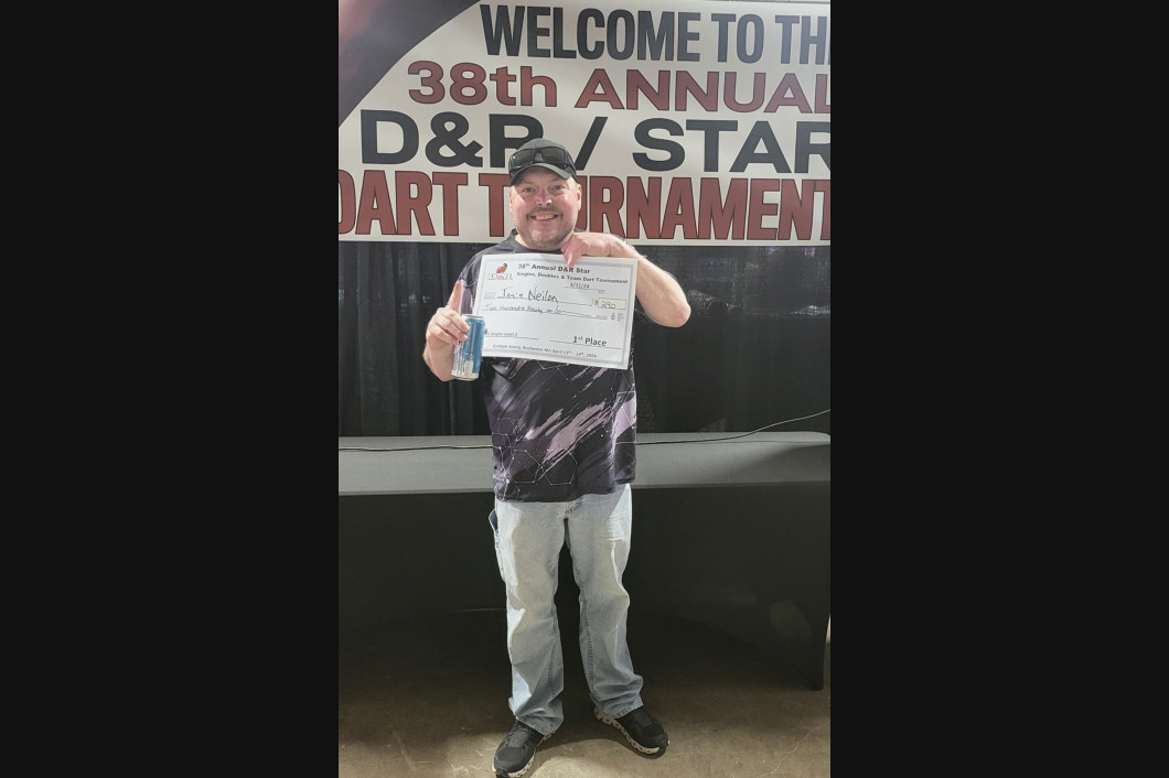 Man holding a paper at a dart tournament, wearing a hat, shirt and jeans. Background banner says 