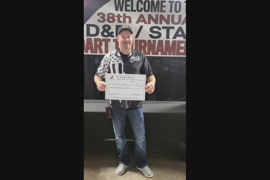 Man holding a check at a dart tournament. He's standing in front of a banner that says 