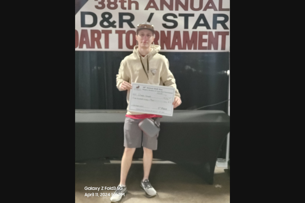 Man in tan hoodie and shorts holds a check, standing in front of a banner for a dart tournament.