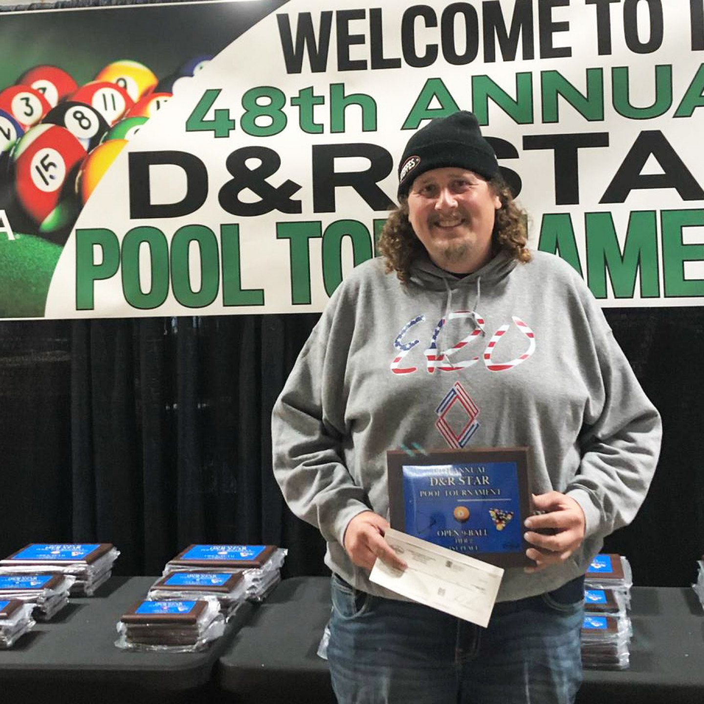 Person holding award and check at a pool tournament.  They wear a hat and gray sweatshirt in front of a banner.