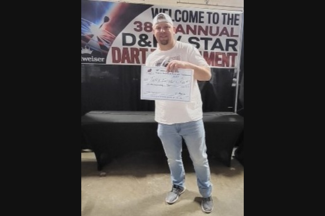 Man holding a document at a darts tournament; standing in front of a banner.