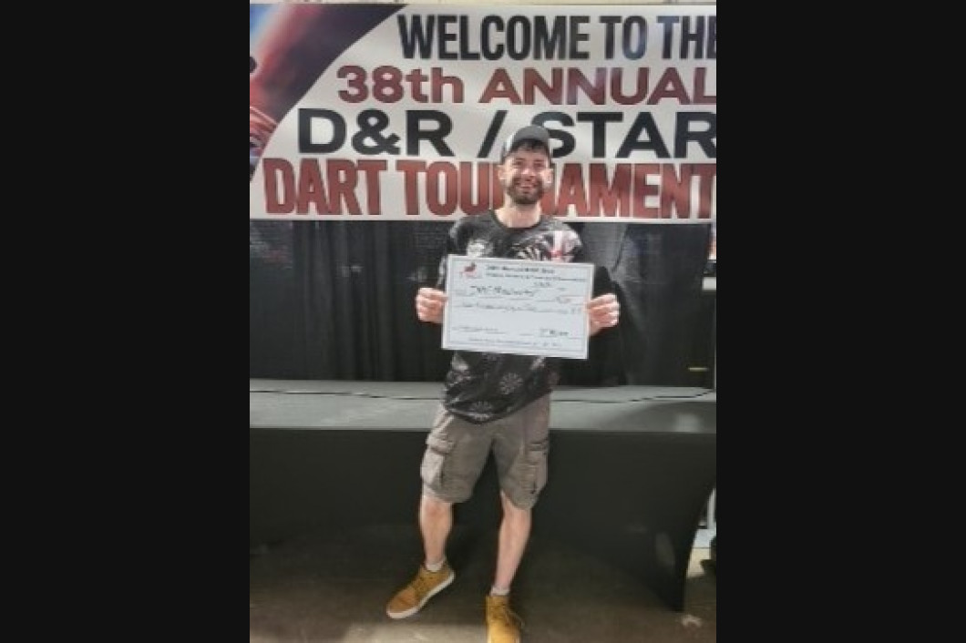 Man holding a certificate at the 38th Annual D&R/Star Dart Tournament. Black backdrop with banner.