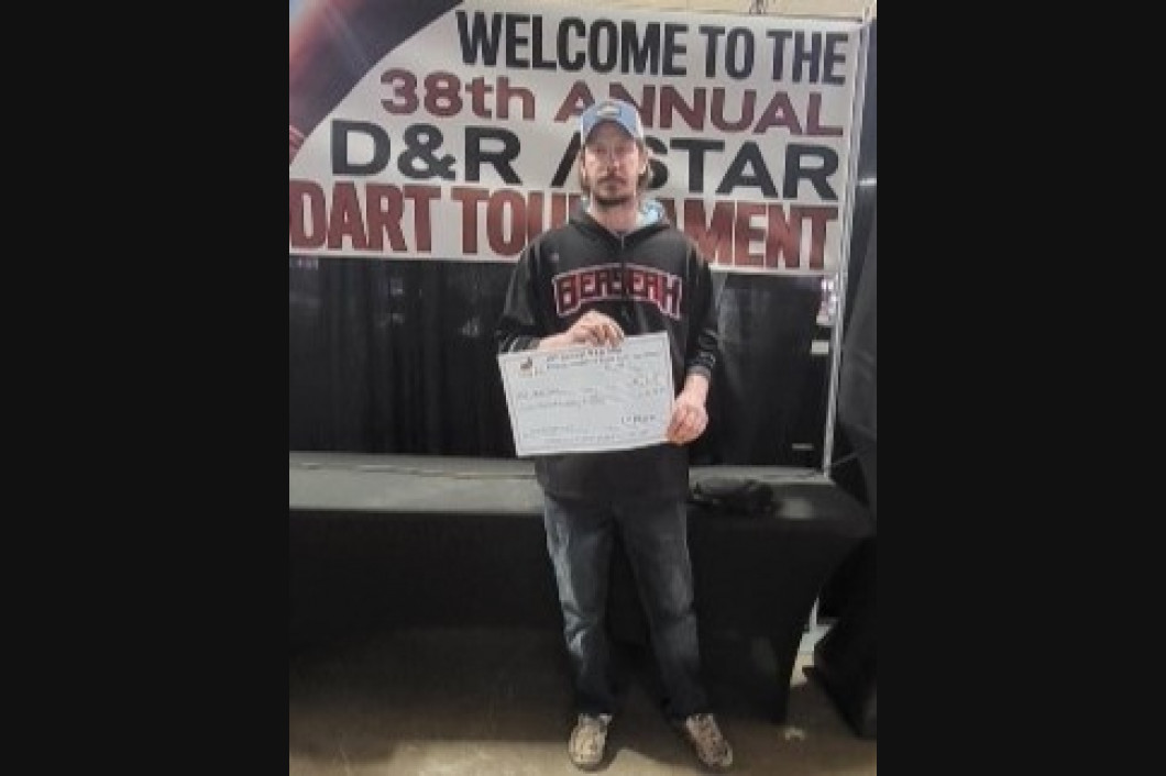 Man in Boston sweatshirt holding a paper, standing in front of a banner for a dart tournament.