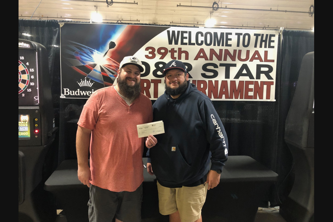 Two men smiling, holding a prize check, at a tournament, in front of a banner that reads 