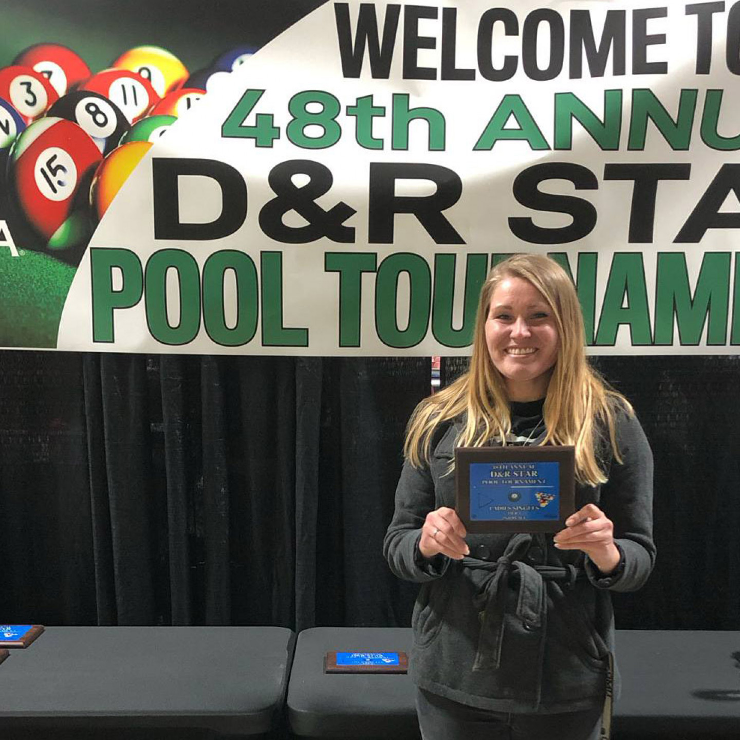 Woman holding award, smiling, in front of a banner for a pool tournament.