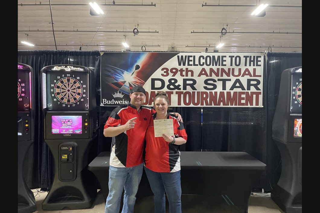 Two people holding certificate, smiling, in front of a darts tournament banner and dart machines.