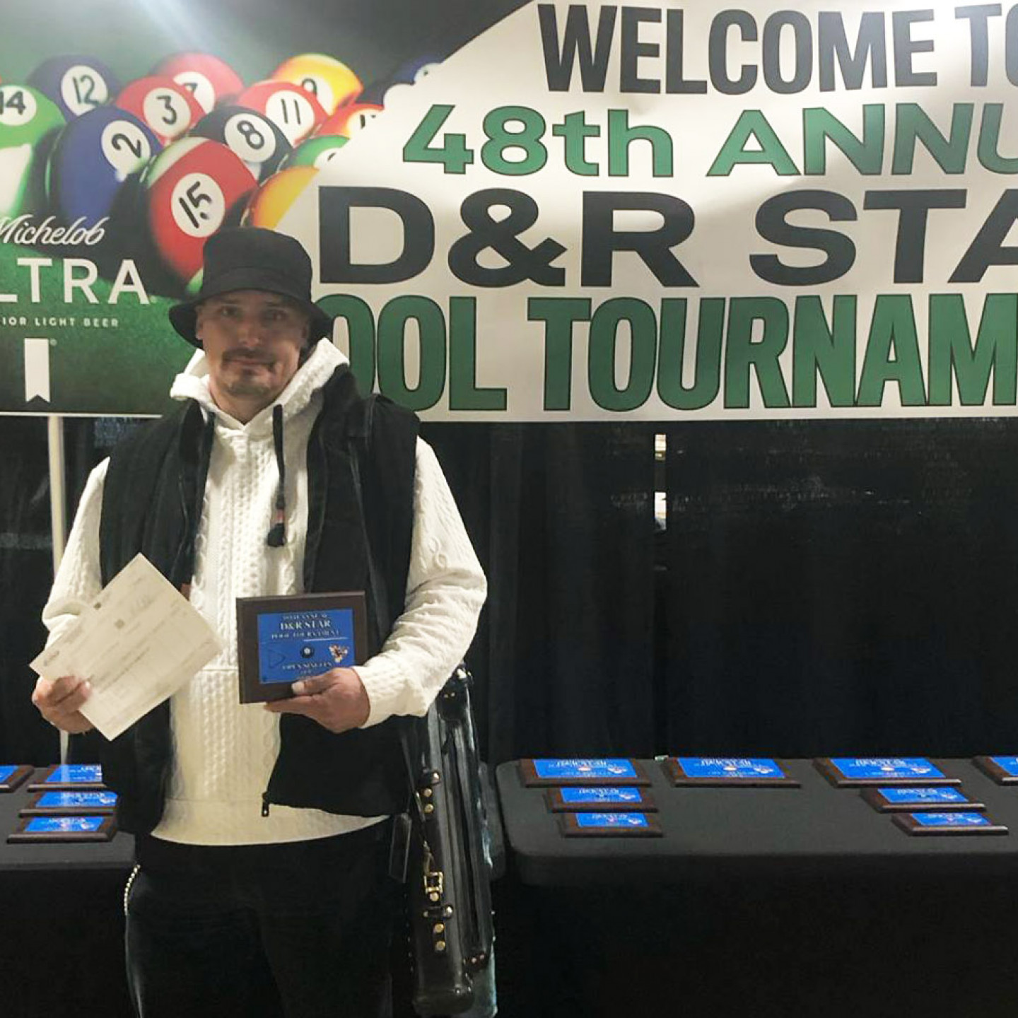 Man in hat holds plaque and papers, at the D&R Star Pool Tournament. Sign reads 