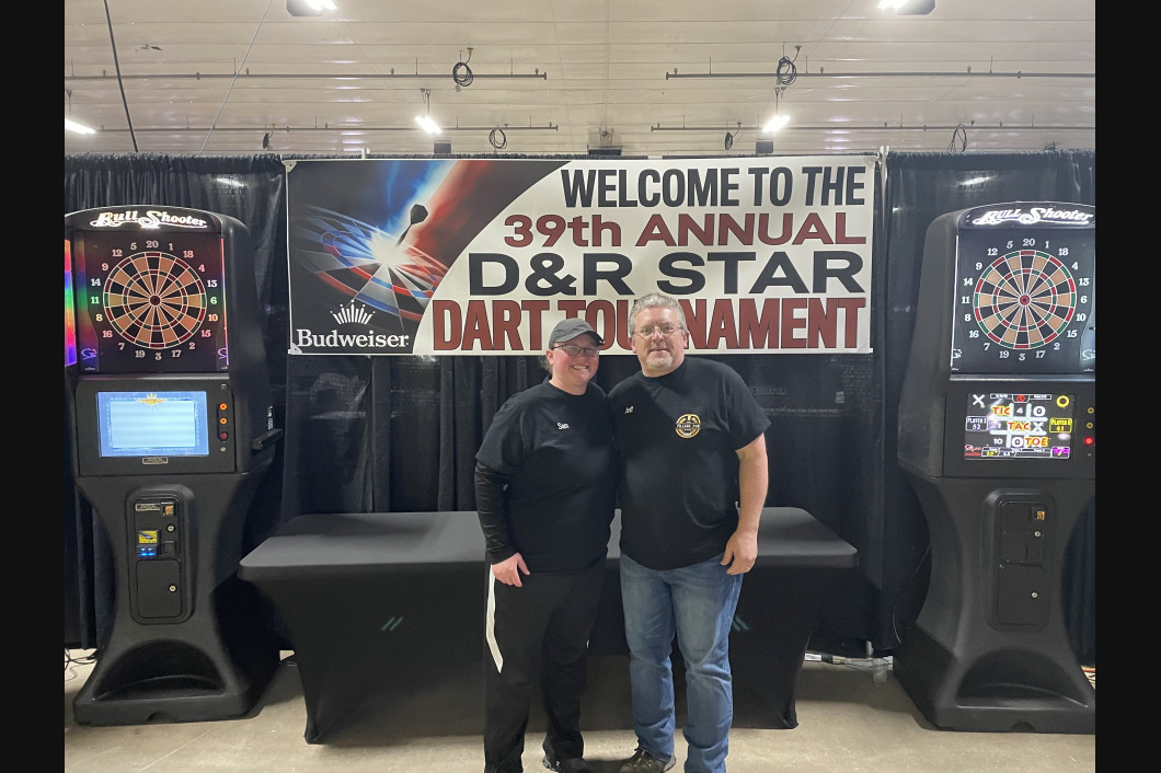 Two people stand in front of dart machines at a tournament. Banner reads: 
