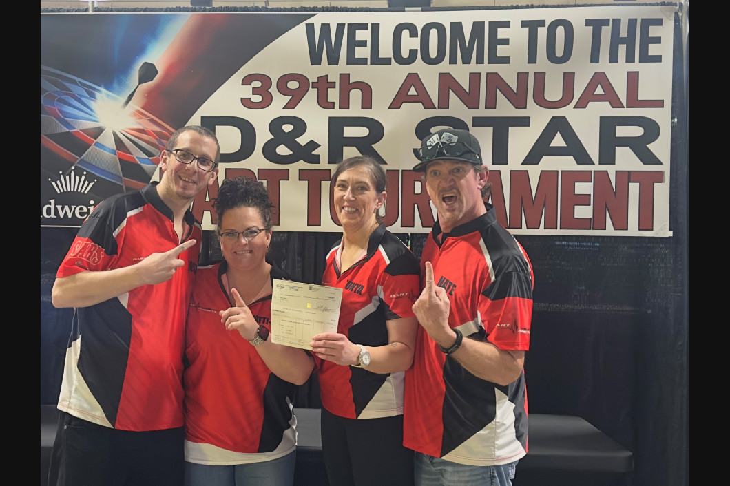 Four people in red/black shirts with trophy, pointing, at 39th D&R Star Dart Tournament banner.