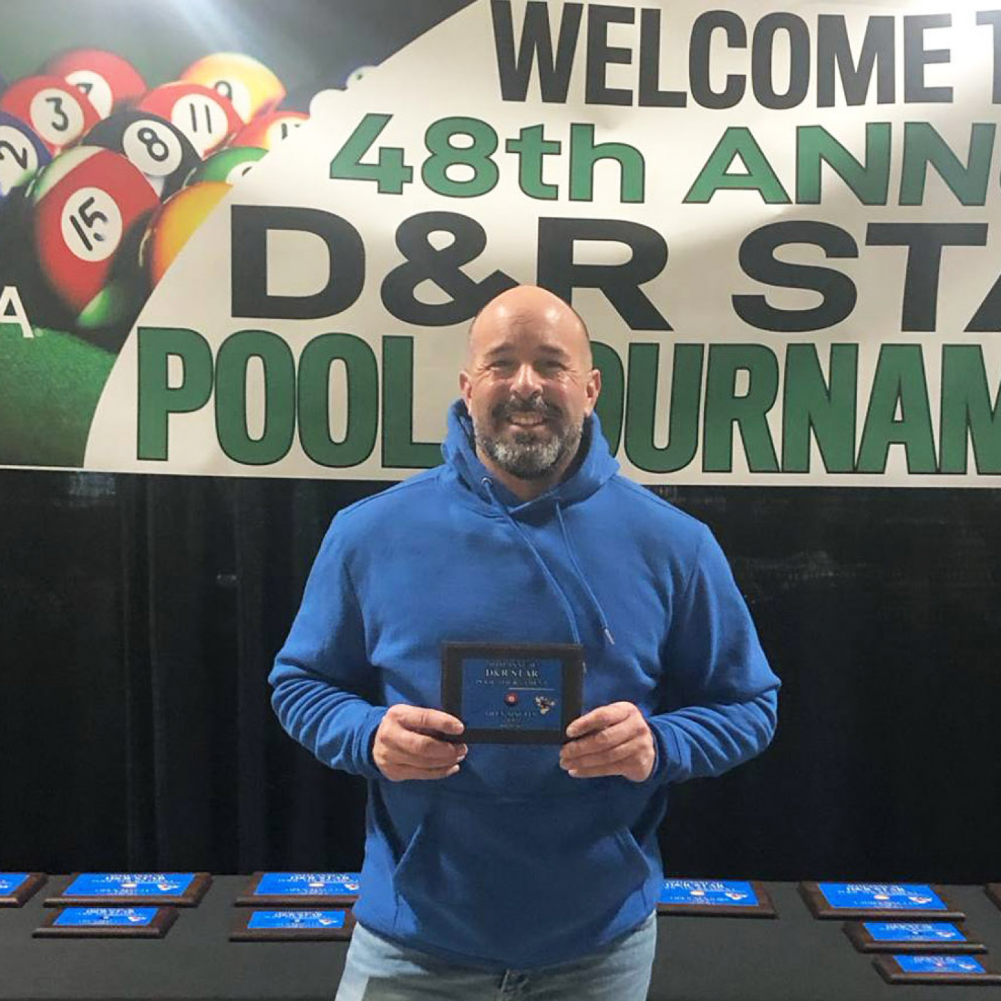 Man in blue hoodie holding a plaque, in front of a pool tournament banner, smiling.