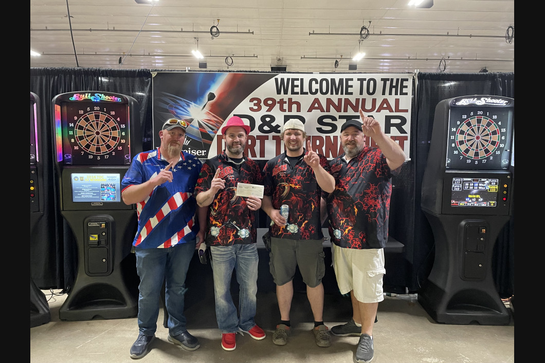 Four men holding a check at a dart tournament. Two dart machines flank them. Banner reads 