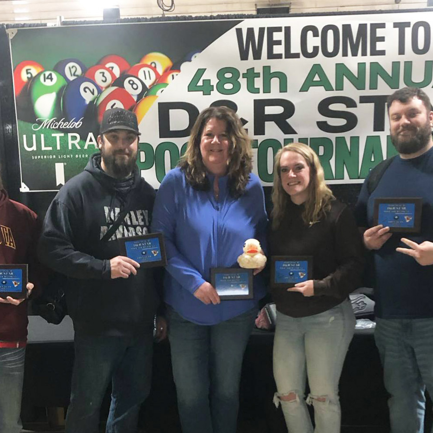 People holding trophies, smiling, standing in front of a tournament banner, and a pool table graphic.