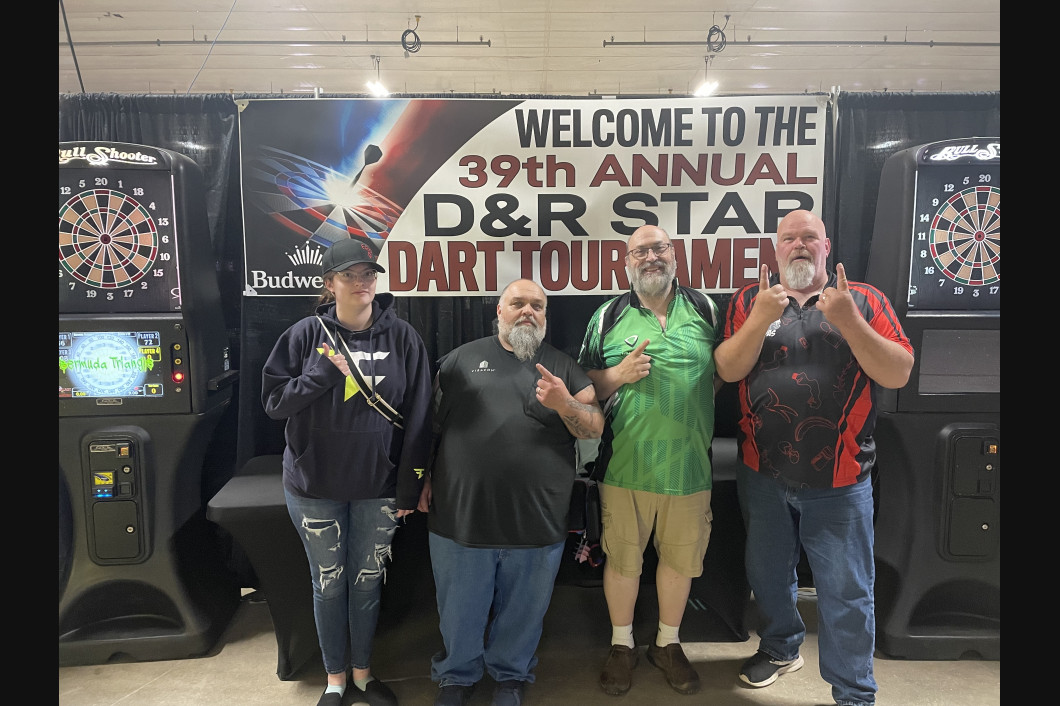 Four people pose in front of a banner for a dart tournament. They give thumbs up.