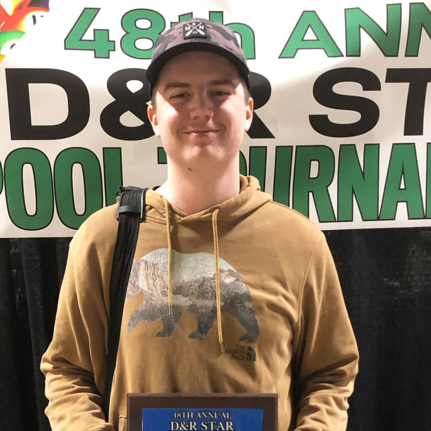 Man in hat and brown hoodie holding an award, standing in front of a banner that reads 
