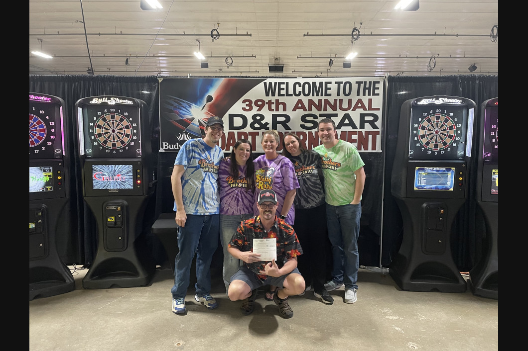 Group of people pose with a certificate in front of dart machines at the 39th annual D&R Star Dart Tournament.