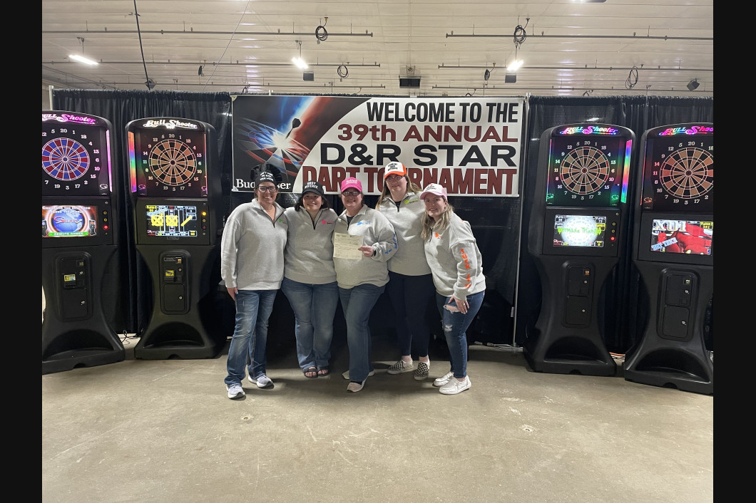 Group of six people smiling, holding a trophy, posing by D&R Star Dart Tournament machines.