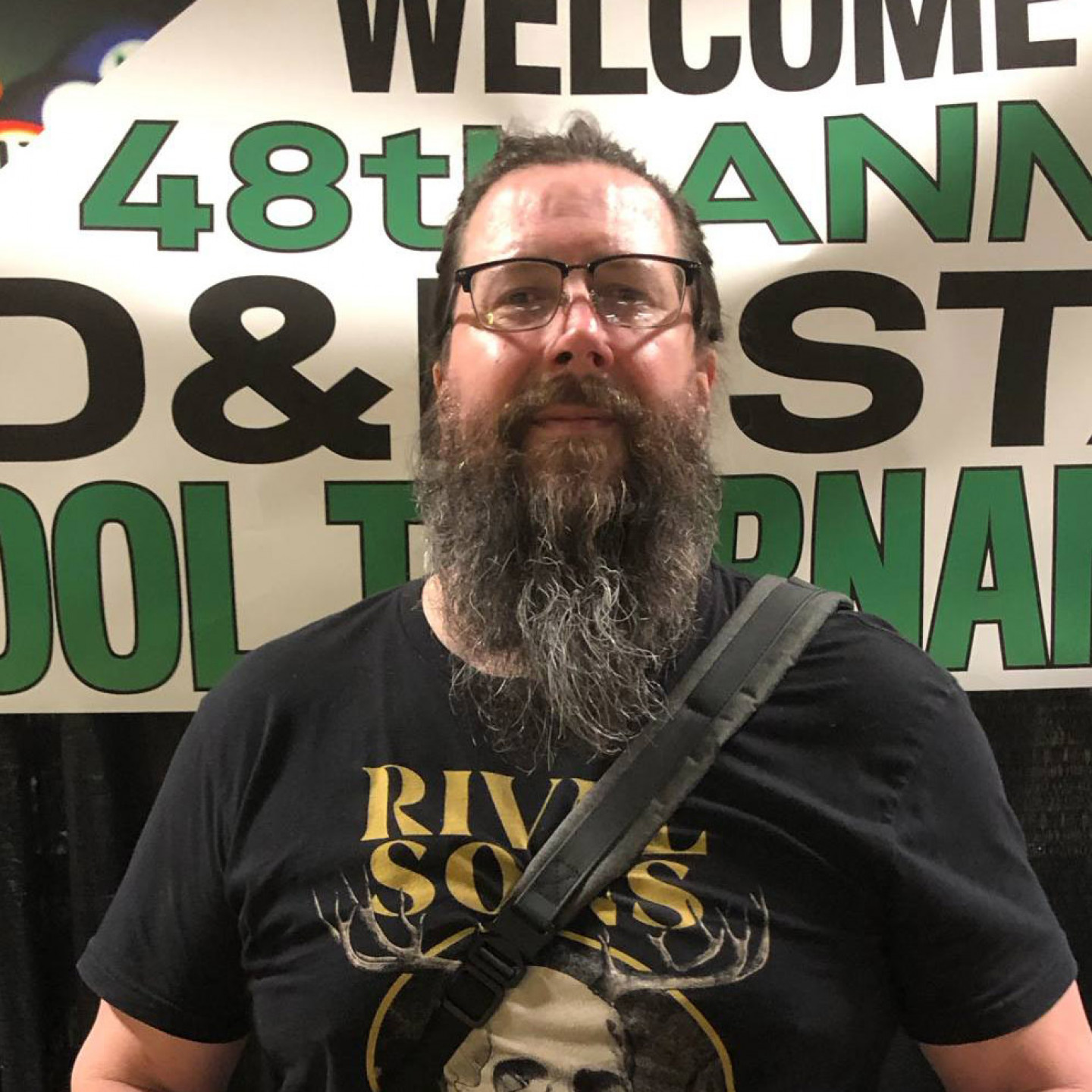 Man with beard, glasses, and a black shirt with text, in front of a banner that reads 
