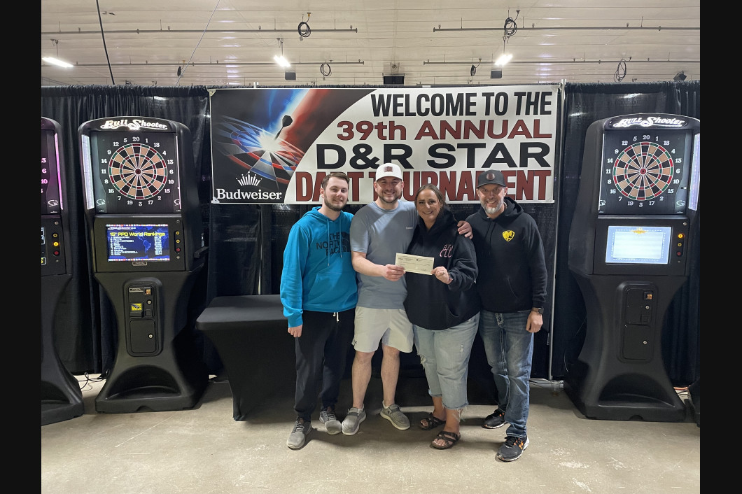 Four people holding a check in front of dart machines at the 39th annual D&R Star Dart Tournament.