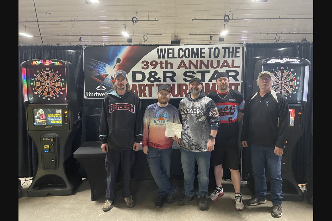 Five people stand in front of dart machines and a tournament banner. They are holding an award, smiling.