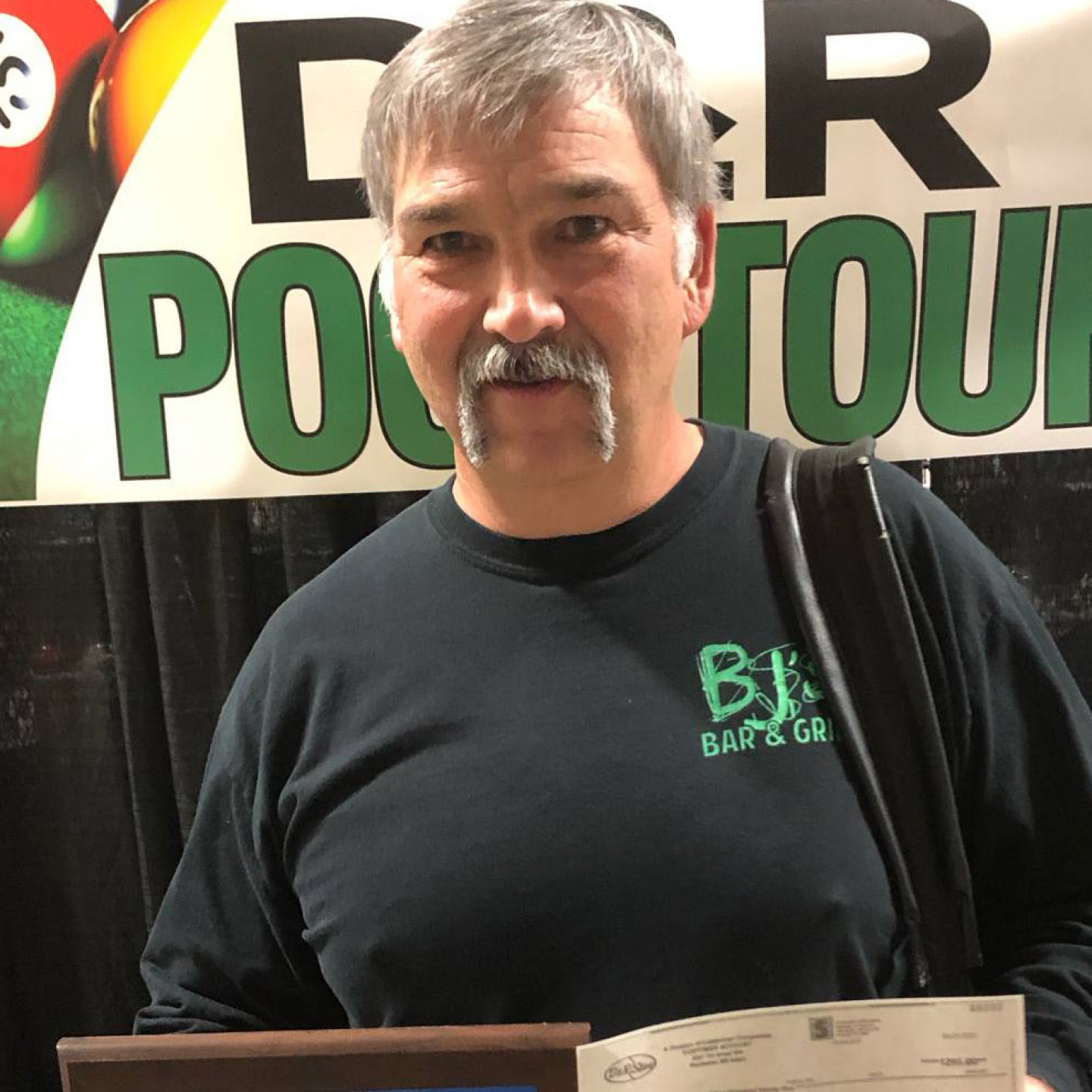 Man in black shirt with mustache holds a trophy and bag, standing in front of a banner that says 