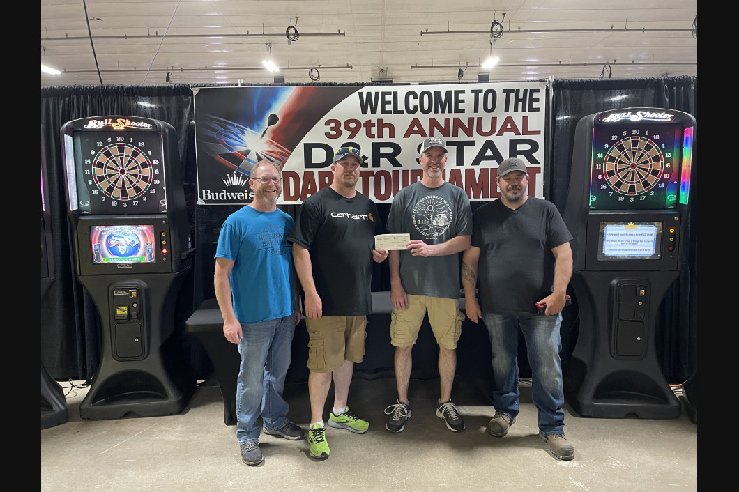 Four men holding a check at a dart tournament. Darts machines behind them; a banner reads “D&R STAR DART TOURNAMENT.