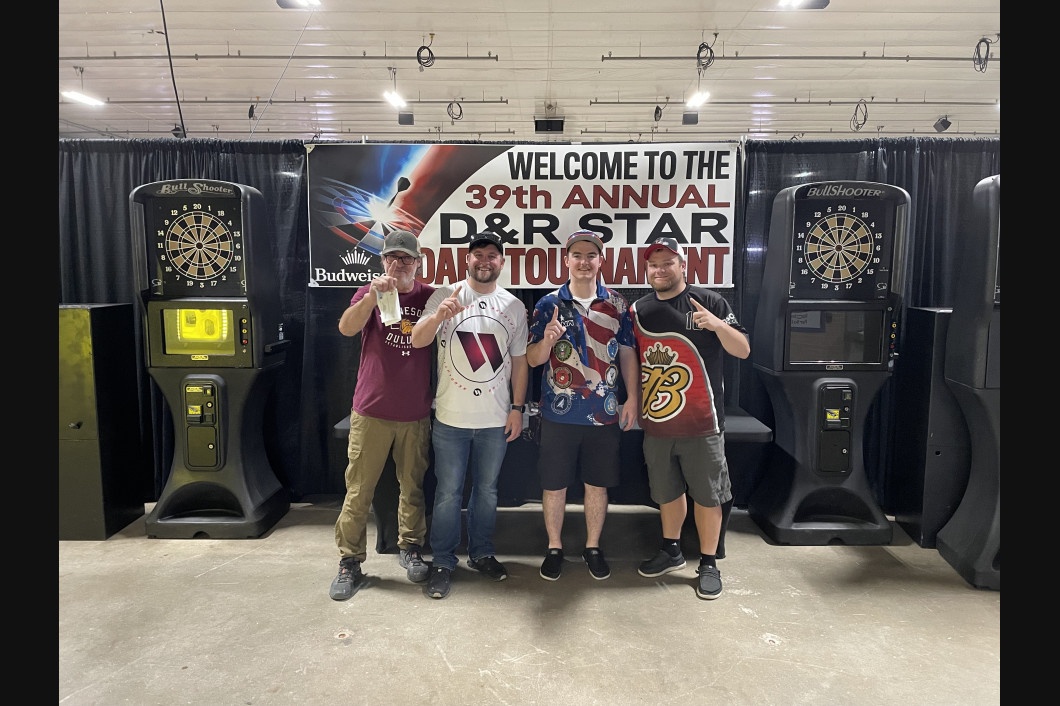Four people posing next to dart machines at a tournament. The sign says “D&R Star Darts Tournament.”