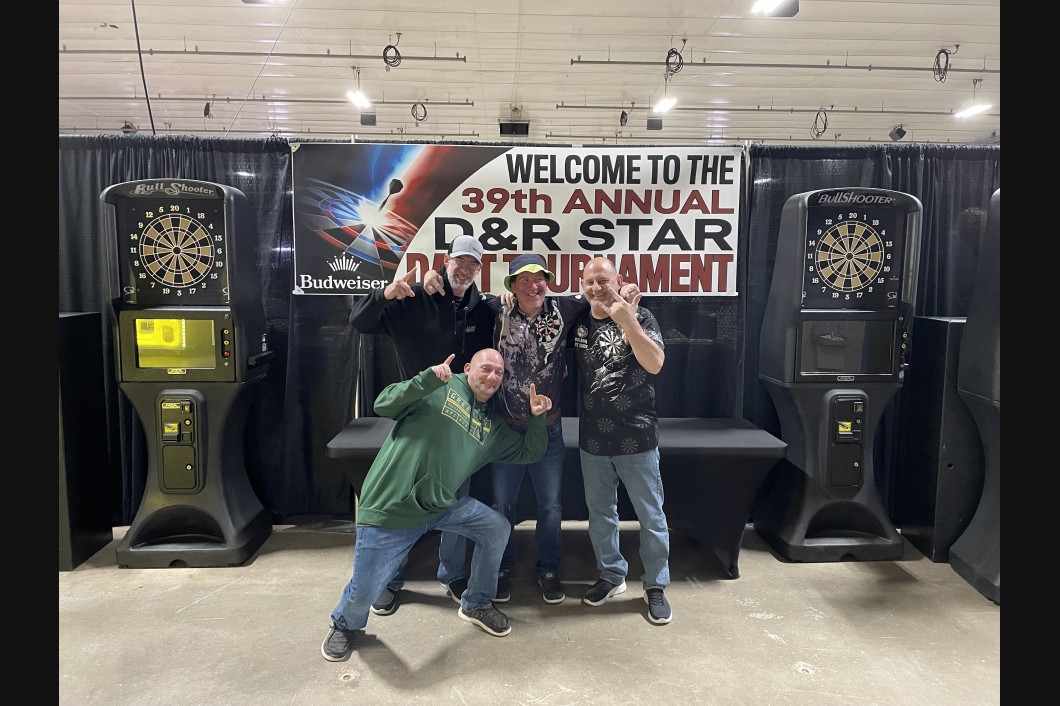 Four men pose in front of a banner for a dart tournament. Two dart machines flank the group.