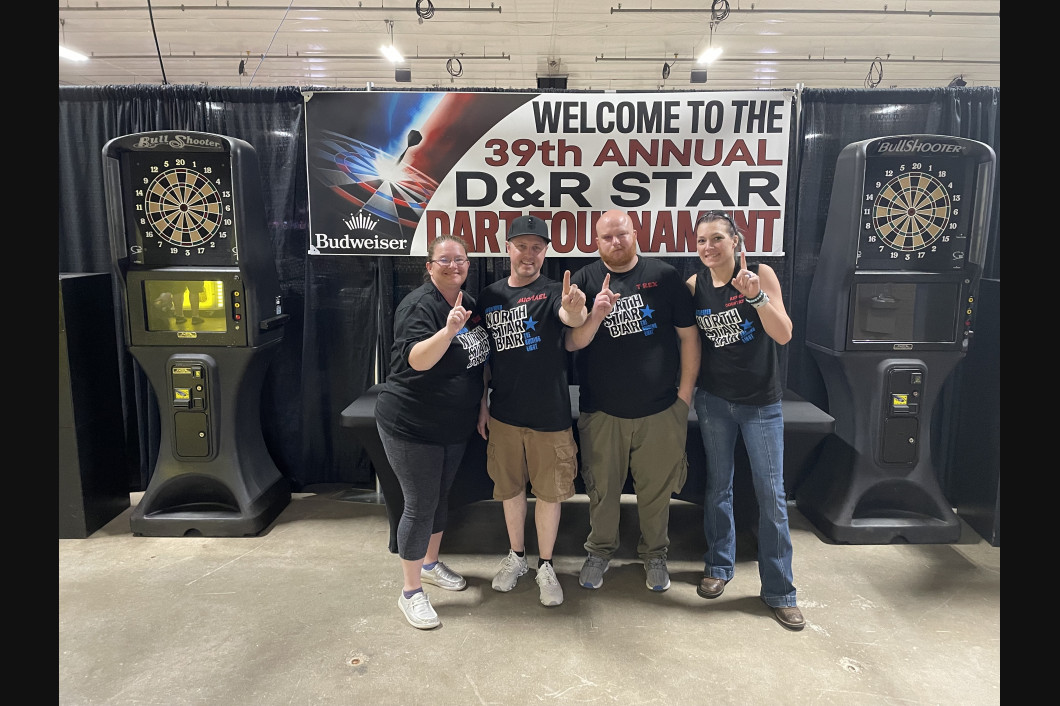 Four people pose in front of dart machines and a tournament banner, all giving thumbs up.