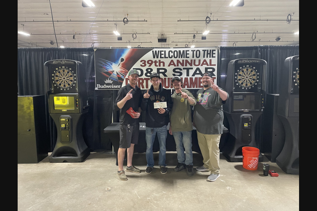 Four people holding a check in front of dart machines at the 